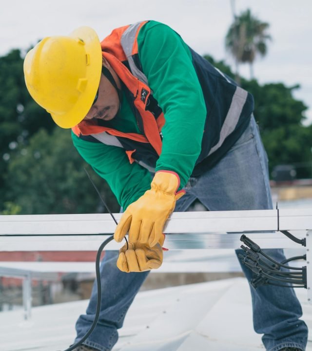 A construction worker wearing PPE installs electrical equipment on a roof.